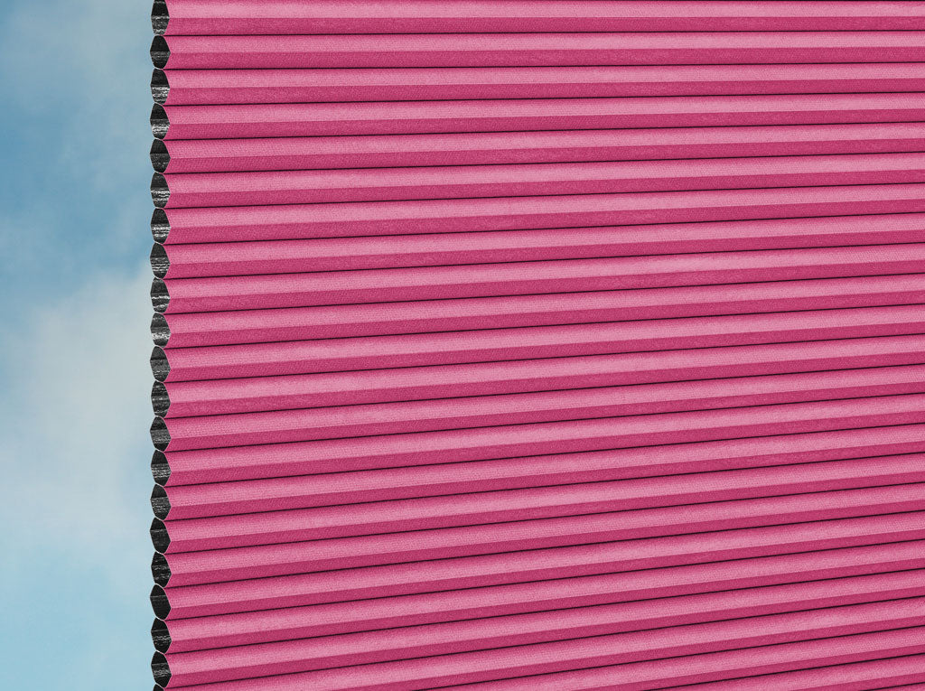 Eine Nahaufnahme der DUETTE® Fixé Unix Dark (3.7493) Rosa Fensterjalousien von Duetteshop zeigt die rechte Seite in Rosa, wobei die linke Kante eine Wabenstruktur aufweist und im Hintergrund ein blauer Himmel mit Wolken zu sehen ist.