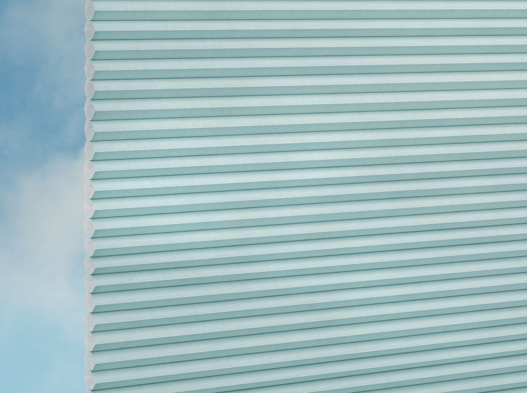 Detailansicht der DUETTE® Fixé Elan (2.7703) Türkis Wabenrollos von Duetteshop mit hellblauem Plisseestoff, teilweise vor einem sanft verschwommenen blauen und weißen Hintergrund dargestellt.