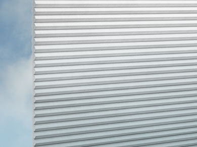 Eine Nahaufnahme von DUETTE® Gradient (3.0802) Türkis von Duetteshop zeigt die weiße, plisseeähnliche, geriffelte Oberfläche mit horizontalen Rillen vor einem blauen Himmel mit weichen Wolken im Hintergrund.
