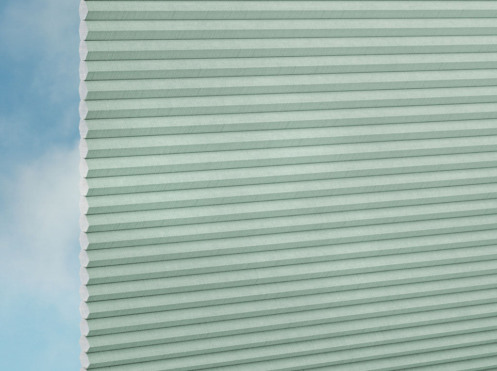 Nahaufnahme der DUETTE® Fixé Batiste (4.6073) Grün Zellulosetöne von Duetteshop, teilweise ausgezogen vor einem unscharfen blau-weißen Himmelshintergrund. Die sechseckige Zellstruktur ist entlang der linken Kante sichtbar.