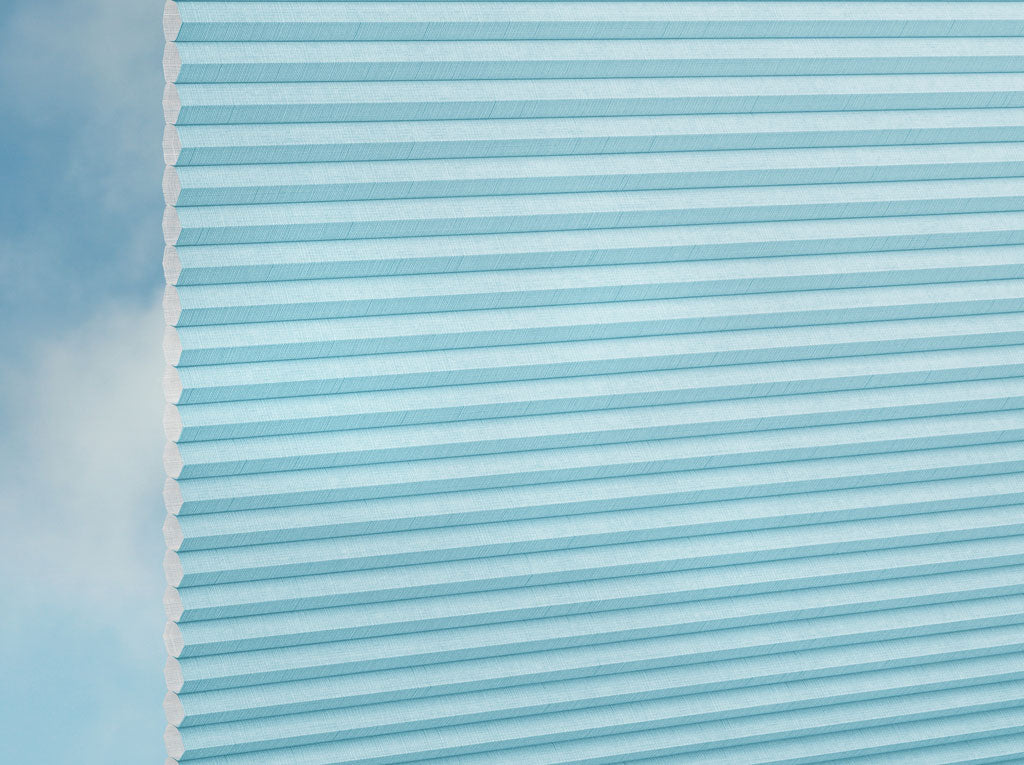 Nahaufnahme des DUETTE® Fixé Batiste (4.6042) Türkis Plissees von Duetteshop mit einer Wabenstruktur in Hellblau vor einem sanft verschwommenen, himmelähnlichen Hintergrund mit blauen und weißen Nuancen.