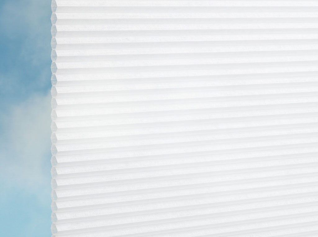 Eine Nahaufnahme von DUETTE® Fixé Batiste (4.3702) Weiß von Duetteshop zeigt die weißen Plissee nach Maß Jalousien teilweise geöffnet, mit sanftem Tageslicht und einem blauen Himmel mit leichten Wolken, die eine helle, luftige Atmosphäre schaffen.
