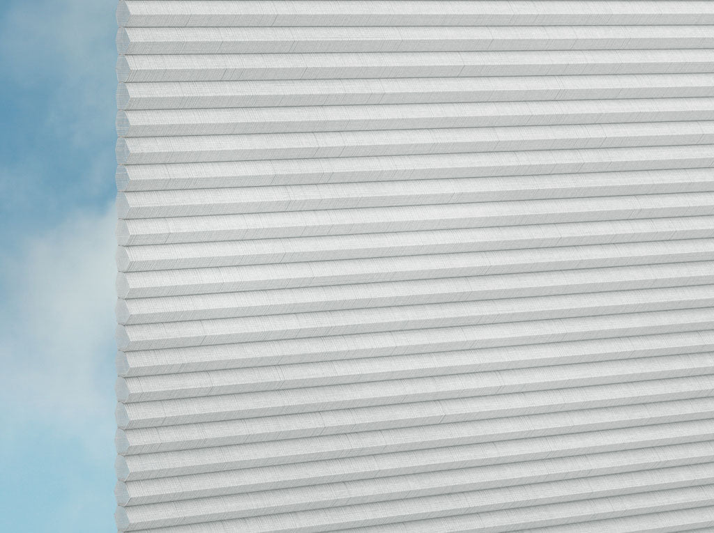 Nahaufnahme von DUETTE® Fixé Batiste (4.3352) Weiß von Duetteshop, mit weißer Wabenstruktur und eleganten Falten. Ein blassblauer Himmel und Wolken im Hintergrund unterstreichen den raffinierten Look dieser maßgefertigten Fensterbekleidung.
