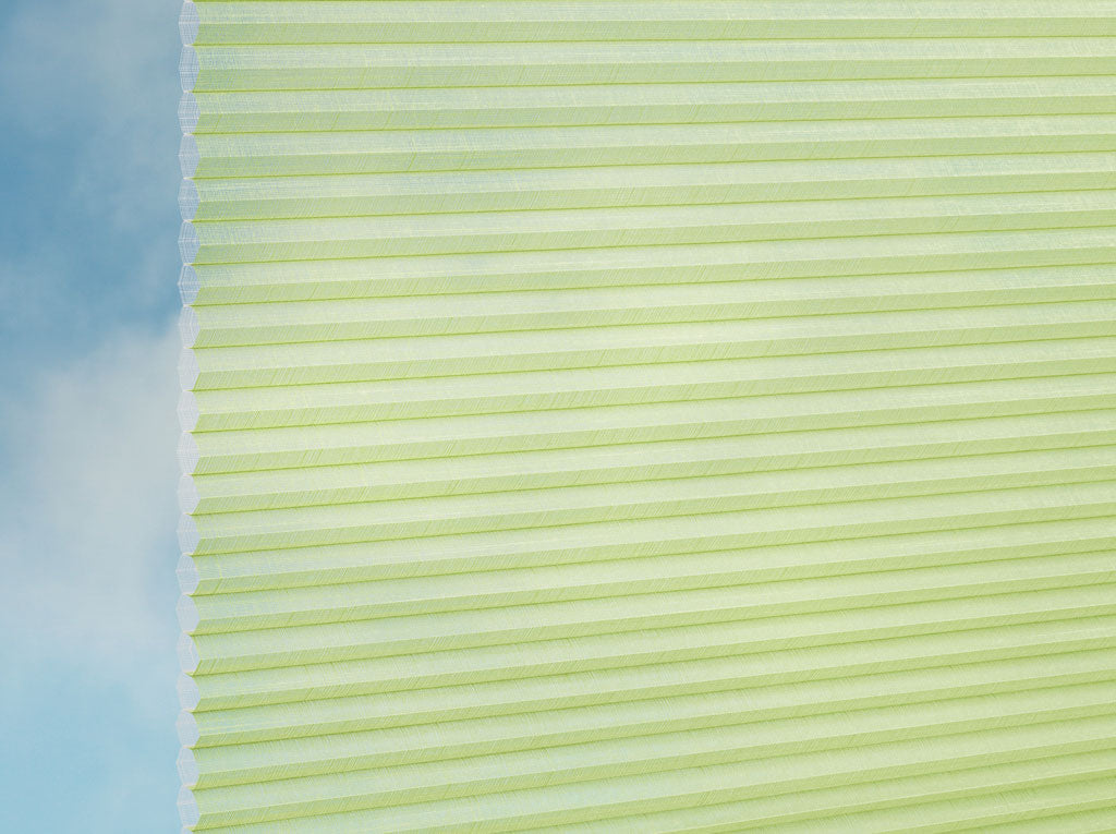 Eine Nahaufnahme des DUETTE® Fixé Batiste Sheer (4.9375) Grün von Duetteshop, die die blassgrünen Falten und die Wabenstruktur zeigt, die das Licht sanft filtern, während links der blaue Himmel zu sehen ist.