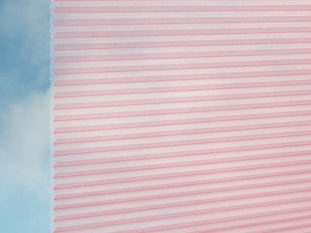 Nahaufnahme des DUETTE® Fixé Batiste Sheer (4.9397) Rosa von Duetteshop, ein hellrosa Plissee mit Wabenstruktur, das einen weichen, wolkenblauen Himmel im Hintergrund teilweise verdeckt.