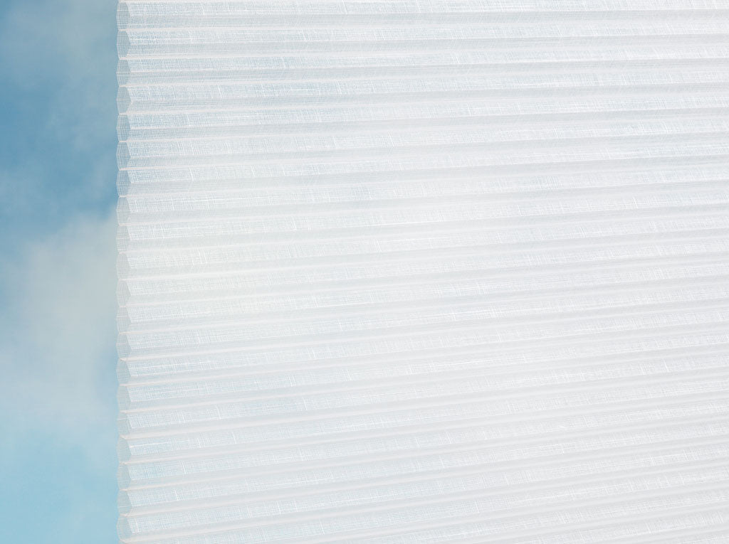 DUETTE® Fixé Batiste Sheer (4.6502) Weiß von Duetteshop besteht aus weißen, lichtdurchlässigen Wabenschirmen, die bei teilweiser Öffnung weiches natürliches Licht filtern. Ein blauer Himmel mit leichten Wolken im Hintergrund unterstreicht die luftige Atmosphäre.